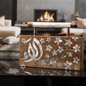 May include: A rectangular wooden clutch bag featuring an inlaid mother-of-pearl floral design on the front panel. The bag includes a top handle with a white shell inlay and dark wood accents, showcasing Arabic calligraphy script in iridescent shell.