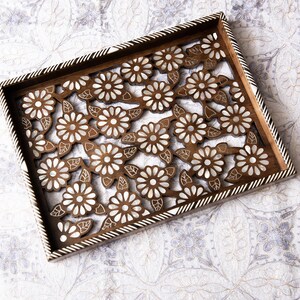 May include: A rectangular wooden tray with a floral cutout design. The tray features a dark brown frame with a white and black striped border. The interior has a repeating pattern of white flowers and leaves against a brown background.