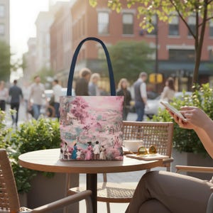 Può includere: Una borsa tote con manico blu navy e stampa di scena pastorale rosa e bianca. La stampa raffigura figure in abiti d'epoca, alberi ed edifici. La borsa è su un tavolo di legno all'aperto.