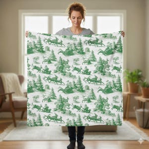 May include: A white blanket with a green toile pattern featuring Santa Claus in a sleigh pulled by reindeer, trees, and other holiday elements. The blanket is held up by a person.