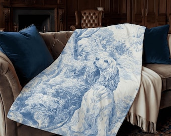 Könnte beinhalten: Eine blau-weiße Decke mit einer detaillierten Illustration eines Hundes in einer Gartenszene. Die Decke ist über ein braunes Sofa mit blauen Kissen drapiert. Der Hund hat eine hellbraune Farbe.
