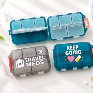 May include: Two rectangular plastic pill organisers with multiple compartments. One is dark blue with the text KEEP GOING, a pink heart, and a yellow pill. The other is translucent grey with the text TRAVEL MEDS, a suitcase icon, and an airplane.