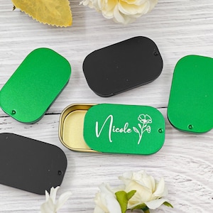 Può includere: Un assortimento di piccole scatole rettangolari in metallo verde e nero. Una scatola verde è aperta, rivelando un interno dorato e il nome "Nicole" con un motivo floreale. Le scatole sono disposte su una superficie di legno chiaro.
