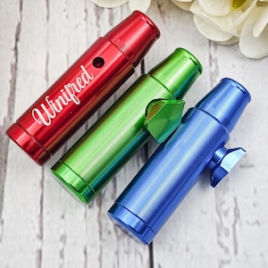 Puede incluir: Tres silbatos metálicos de colores en rojo, verde y azul. El silbato rojo tiene el nombre "Winifred" en escritura blanca. Los silbatos son cilíndricos con una textura acanalada y un pequeño agujero o palanca.