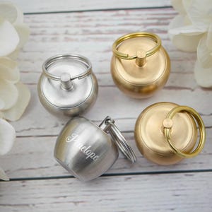 Puede incluir: Cuatro llaveros metálicos en tonos plateados y dorados. Un llavero plateado está grabado con el nombre "Penelope". Los llaveros tienen forma redonda con un anillo para llaves. El fondo incluye flores blancas y una superficie de madera.