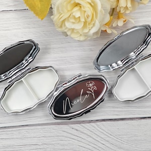 May include: Oval-shaped pill cases with silver-colored trim and a two-compartment interior. One case has the name "Nicole" and a floral design engraved on the lid. The cases are open and closed, showcasing the interior and exterior.