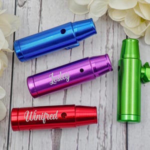 Puede incluir: Vista superior de cuatro artículos metálicos personalizados. Tres son cilíndricos, en azul, morado y rojo, cada uno con un nombre grabado. El cuarto es verde, con una perilla. Sobre una superficie de vetas de madera clara.