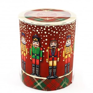 May include: A cylindrical candle holder with a red background and white star accents. The holder features four colourful nutcracker soldier illustrations. The top and bottom have a red and green plaid pattern. The lid reads "Christmas Magic".