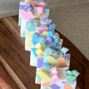 May include: Artisan soap bars with a white base, adorned with colorful, geometric shapes and heart accents in pastel shades of pink, blue, yellow, and purple. The soaps are arranged on a wooden surface.