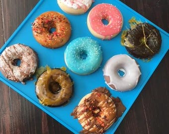 May include: A collection of nine donuts with various toppings and glazes, arranged on a bright blue square tray. The donuts feature sprinkles, chocolate, and pink and white frosting. The background is a dark wooden surface.