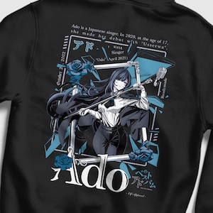 May include: A black hoodie featuring a graphic print of a long-haired character holding a microphone inside a frame. The design includes blue roses, the text Ado, and biographical details about the Japanese singer in white lettering.