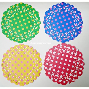 Könnte beinhalten: Vier Papierdeckchen mit einem Spitzenmuster und Punkten in verschiedenen Farben. Die Deckchen sind grün mit gelben Punkten, blau mit rosa Punkten, gelb mit rosa Punkten und rot mit weißen Punkten.