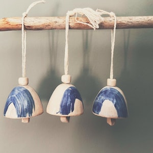 May include: Three ceramic bells with a cream base and blue painted accents hang from a wooden branch. Each bell is suspended by a white string and has a small wooden bead at the top. The bells are a decorative item.