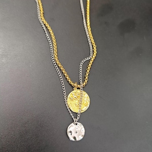 Könnte beinhalten: Eine mehrreihige Halskette mit zwei Ketten, eine goldfarbene und eine silberfarbene. Die goldfarbene Kette hält einen goldenen Anhänger mit detailliertem Design, während die silberfarbene Kette einen kleineren, silbernen Anhänger mit strukturierter Oberfläche hat. Die Halsketten werden auf dunklem Untergrund gezeigt.