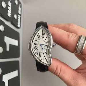 May include: A wristwatch with an asymmetrical, distorted silver-tone case and a white dial featuring elongated black Roman numerals. The watch has thin blue hands and a black leather strap. A silver-tone ring with a textured finish is also visible.