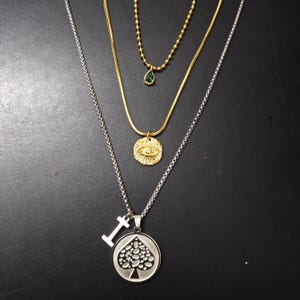 Könnte beinhalten: Eine Sammlung von drei Halsketten. Die oberste Halskette ist goldfarben mit einem kleinen grünen Tropfenanhänger. Die mittlere Halskette ist goldfarben mit einem Augenanhänger. Die untere Halskette ist silberfarben mit einem Pik-Anhänger.