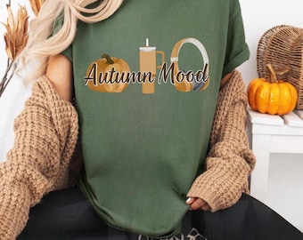 Könnte beinhalten: Ein olivgrünes T-Shirt mit einem Grafikdruck, der einen Kürbis, einen hohen Becher mit Strohhalm und einen Kopfhörer zeigt. Der Text "Autumn Mood" ist in einer kursiven braunen Schrift in der Mitte des Grafikdesigns auf dem T-Shirt gedruckt.