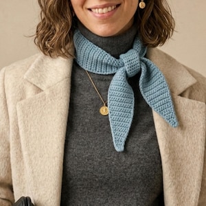 Op de afbeelding: Een lichtblauwe gehaakte sjaal, om de nek geknoopt. De sjaal wordt gecombineerd met een grijze coltrui en een beige jas. Een gouden hangerketting en oorbellen voegen een vleugje elegantie toe aan de outfit.