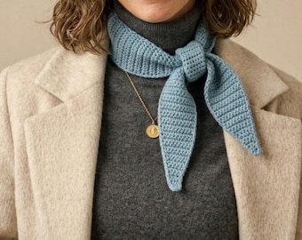 Può includere: Sciarpa all'uncinetto azzurra annodata al collo. È abbinata a un dolcevita grigio e a un cappotto beige. Una collana con pendente dorato e orecchini aggiungono un tocco di eleganza all'outfit.