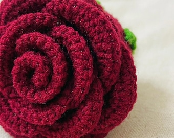 Può includere: Una rosa all'uncinetto color bordeaux intenso con petali strettamente arrotolati e una piccola foglia verde visibile sul lato. La trama del filato è densa e uniforme, creando una forma floreale arrotondata e stratificata con un sottile riflesso.