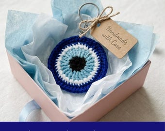 Può includere: Un portachiavi all'uncinetto a forma di occhio blu, bianco e nero, presentato in una scatola regalo rosa con carta velina blu. Un piccolo tag legato con spago recita "Handmade with Care". Il testo "Crochet Bookmark Pattern PDF" è in basso.