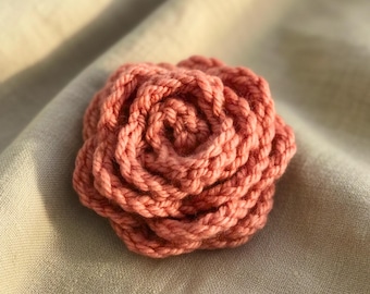 May include: A handmade, coral-colored crocheted rose. The flower is in full bloom, with tightly wound petals creating a textured, three-dimensional effect. The rose is set against a neutral, light beige fabric background.