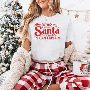 May include: White t-shirt with the text "DEAR Santa I CAN EXPLAIN" in red. The image also includes a red and white plaid pajama set and a mug with a Santa Claus design. The image is festive and holiday-themed.