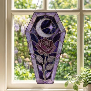 May include: A coffin-shaped stained glass suncatcher featuring a crescent moon and a four-pointed star at the top, with a central rose and leaves below. The piece is constructed from various shades of purple and violet glass with dark metal cames.