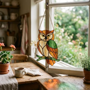 May include: A stained glass suncatcher shaped like an owl perched on a branch. The owl features amber, brown, cream, and green glass panels held together by dark metal lines. It hangs from a gold-toned metal chain attached to a window frame.