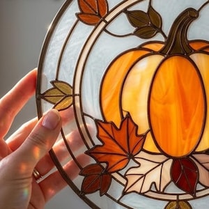 May include: A circular stained glass suncatcher featuring a central orange pumpkin surrounded by various autumn leaves in shades of amber, brown, and cream. The design is encased in a dark metal frame with a textured, translucent white glass background.