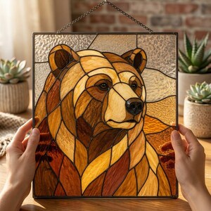 Può includere: Un pannello quadrato in vetro colorato che presenta il ritratto dettagliato di un orso bruno. L'orso è composto da vari pezzi di vetro color ambra, marrone chiaro e scuro uniti da linee metalliche scure. Lo sfondo include vetro trasparente testurizzato.