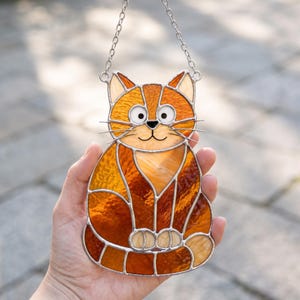 Könnte beinhalten: Ein Buntglas-Sonnenfänger in Form einer sitzenden Katze mit strukturierten orangefarbenen und bernsteinfarbenen Glasscheiben. Die Katze hat weiße Glasaugen, eine schwarze Metallumrandung und eine silberne Kette zum Aufhängen.