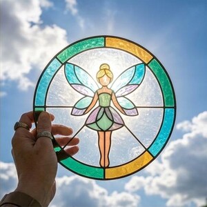 May include: A circular stained glass suncatcher featuring a central fairy figure with translucent wings. The design includes a green and yellow outer border, with the fairy rendered in shades of light green, purple, and clear glass segments.
