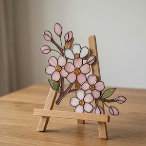 May include: A stained glass floral piece featuring pink and white blossoms with yellow centres and green leaves, supported by a light-coloured wooden easel. The glass petals are outlined in dark metal, creating a delicate botanical arrangement.