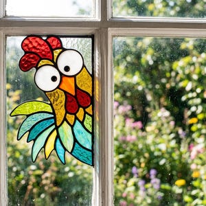 May include: A stained glass suncatcher shaped like a rooster head with large white eyes, a bright orange beak, and a red comb. The rooster features textured yellow, teal, and light blue glass pieces, all outlined in black metal solder lines.