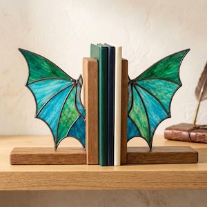 May include: A pair of wooden bookends featuring stained glass wings in shades of teal, turquoise, and emerald green. The wings are constructed with dark metal soldering and are attached to rectangular wooden bases, holding three upright books.
