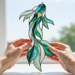 May include: A stained glass suncatcher shaped like a koi fish, featuring various shades of emerald, teal, and white glass pieces joined by dark metal solder lines. The fish has a small gold-coloured accent near the head and a hanging loop.