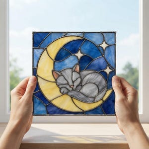 May include: A square stained glass panel featuring a gray cat curled up sleeping on a yellow crescent moon. The background consists of various shades of blue glass with small white star shapes. The piece is held up against a bright window.
