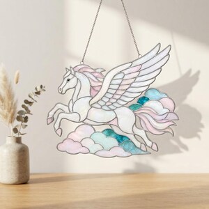 May include: A stained glass suncatcher shaped like a winged horse, or pegasus, in flight. The piece features iridescent white, soft pink, and teal glass segments outlined in dark metal, suspended by a thin chain above a cluster of clouds.