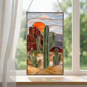 May include: A rectangular stained glass panel featuring a desert landscape with a tall green saguaro cactus, a smaller prickly pear cactus with red accents, brown rock formations, a bright orange sun, and a light blue sky, all held by a metal chain.