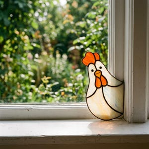 Op de afbeelding: Een glas-in-lood zonnevanger in de vorm van een kip, met een gebroken wit lichaam en een feloranje kam en lellen. Het object is vervaardigd met donkere metalen soldeerlijnen en staat tegen een raamkozijn om het natuurlijke licht op te vangen.