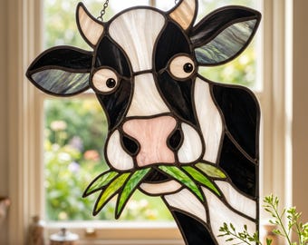 Könnte beinhalten: Ein Buntglas-Suncatcher in Form eines Kuhkopfes mit schwarzen und weißen Glaspaneelen, einer rosa Nase und grünen Glas-Grasbüscheln im Maul. Das Stück weist dunkle Metall-Lötlinien auf und ist an einer dünnen Metallkette aufgehängt.