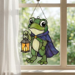 Könnte beinhalten: Ein Buntglas-Sonnenfänger in Form eines grünen Frosches, der einen violetten Umhang trägt und eine kleine Laterne mit warmem gelbem Licht hält. Das Stück hängt an einer Metallkette und hat dunkle Bleilinien, die die Glassegmente umranden.