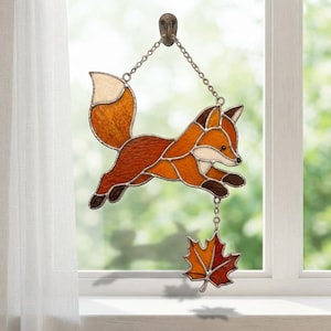 Peut inclure: Un attrape-soleil en vitrail en forme de renard bondissant, composé de segments de verre orange, marron et crème assemblés par des soudures métalliques. Une petite chaîne suspend une feuille d'érable en verre teinté orange et rouge.