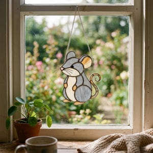 May include: A stained glass suncatcher shaped like a mouse with grey and cream-colored glass panels, black solder lines, and a silver-toned metal chain for hanging. The mouse features a curled tail, rounded ears, and a small pink nose detail.