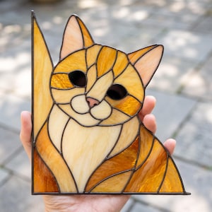 May include: A stained glass suncatcher shaped like an orange tabby cat. The piece features various shades of amber, gold, and cream glass segments held together by dark metal solder lines, forming the head and chest of a cat with pointed ears.