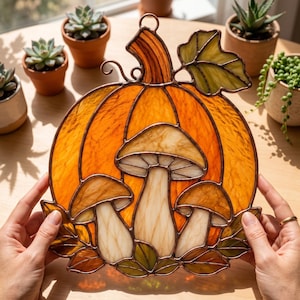 May include: A stained glass suncatcher featuring a large orange pumpkin with a green leaf and three cream-colored mushrooms at the base. The design is outlined in dark metal solder, creating a decorative piece with a hanging loop at the top.