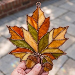Puede incluir: Un atrapasoles de vidrieras con forma de hoja de arce y dos bellotas adjuntas. La hoja presenta segmentos de vidrio ámbar, naranja, amarillo y verde oliva unidos por líneas de soldadura de metal color cobre. Tiene una pequeña anilla arriba.