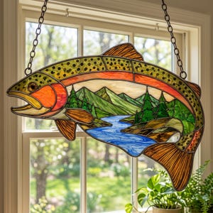 May include: A stained glass suncatcher shaped like a trout, featuring a landscape scene with green mountains, evergreen trees, and a blue river. The fish has a spotted yellow and orange body with dark outlines, suspended by two metal chains.