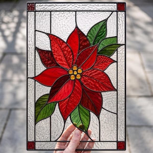 May include: A rectangular stained glass panel featuring a central red poinsettia flower with green leaves and yellow glass beads at the center. The background is made of clear textured glass, and the entire piece is framed in black metal with small red glass accents in each corner.